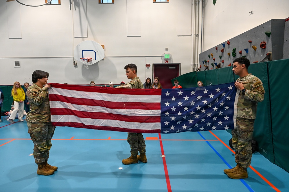 Three military personnel in OCP (Operational Camouflage Pattern) uniforms are holding an unfolded American flag horizontally in a school gymnasium. They appear to be conducting a flag folding demonstration or ceremony for an audience of students visible in the background. The setting includes a basketball hoop, climbing wall, and padded gym walls.
