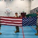 Three military personnel in OCP (Operational Camouflage Pattern) uniforms are holding an unfolded American flag horizontally in a school gymnasium. They appear to be conducting a flag folding demonstration or ceremony for an audience of students visible in the background. The setting includes a basketball hoop, climbing wall, and padded gym walls.
