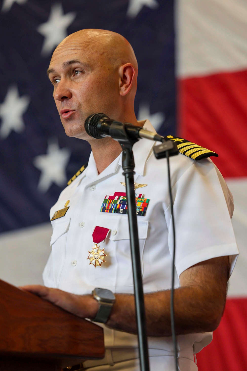 A senior U.S. Navy officer in white service dress uniform is speaking at a podium with a microphone, in front of a large American flag. He is wearing a Legion of Merit medal, aviator wings, multiple service ribbons, and shoulder boards indicating the rank of Captain (O-6). The setting appears to be a formal military ceremony.