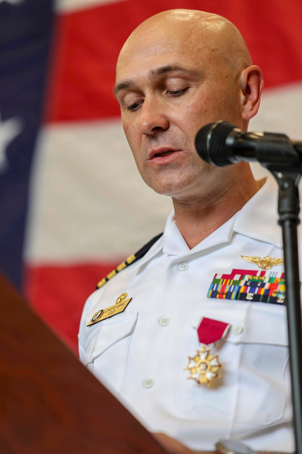 A bald male military officer in a white dress uniform is speaking at a podium with a microphone. He is wearing multiple service ribbons, naval aviator wings, and a prominent medal with a red ribbon on his chest. An American flag is visible in the background.