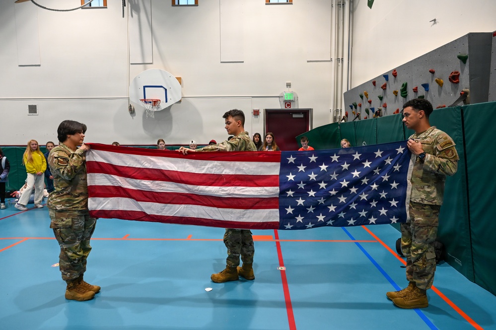 Three military personnel in OCP (Operational Camouflage Pattern) uniforms are holding an unfolded American flag horizontally in a school gymnasium. Students are seated along the wall in the background observing the demonstration. The setting appears to be an educational event, likely a flag folding ceremony demonstration for school children.