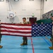 Three military personnel in OCP (Operational Camouflage Pattern) uniforms are holding an unfolded American flag horizontally in a school gymnasium. Students are seated along the wall in the background observing the demonstration. The setting appears to be an educational event, likely a flag folding ceremony demonstration for school children.