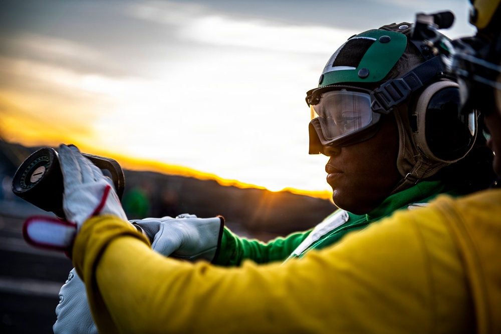 A military or naval aviation deck crew member is photographed at sunset or sunrise, wearing distinctive colored safety gear. The individual is wearing a green helmet with protective goggles, hearing protection, and a yellow jersey, which are standard color-coded uniforms used on aircraft carrier flight decks. They appear to be handling equipment, likely performing pre-flight or maintenance duties.