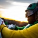 A military or naval aviation deck crew member is photographed at sunset or sunrise, wearing distinctive colored safety gear. The individual is wearing a green helmet with protective goggles, hearing protection, and a yellow jersey, which are standard color-coded uniforms used on aircraft carrier flight decks. They appear to be handling equipment, likely performing pre-flight or maintenance duties.