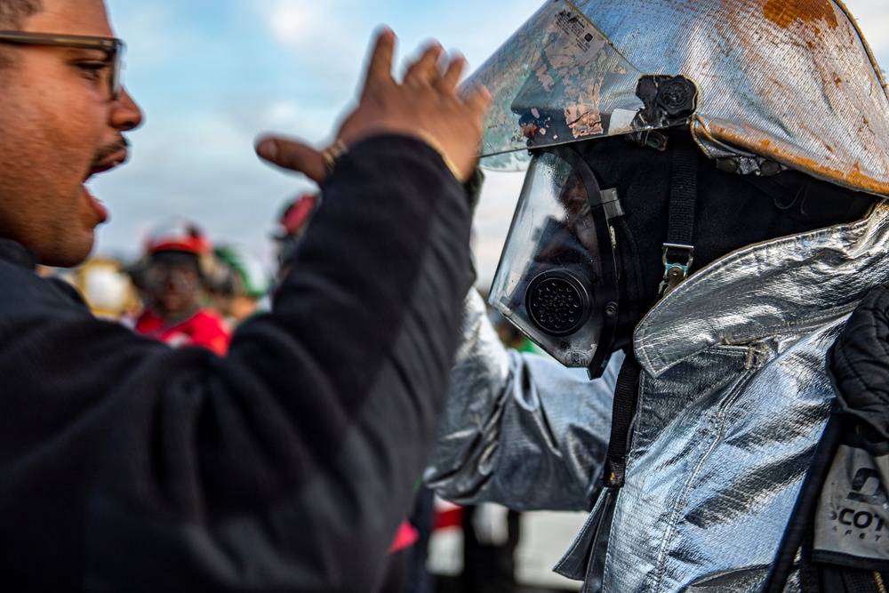 A person in civilian clothing is adjusting or inspecting the visor of a firefighter or hazmat responder wearing a reflective aluminized proximity suit and full-face respirator mask. The protective gear shows signs of wear and heat damage, including scorching on the helmet visor. Additional personnel in safety helmets are visible in the blurred background.