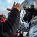 A person in civilian clothing is adjusting or inspecting the visor of a firefighter or hazmat responder wearing a reflective aluminized proximity suit and full-face respirator mask. The protective gear shows signs of wear and heat damage, including scorching on the helmet visor. Additional personnel in safety helmets are visible in the blurred background.