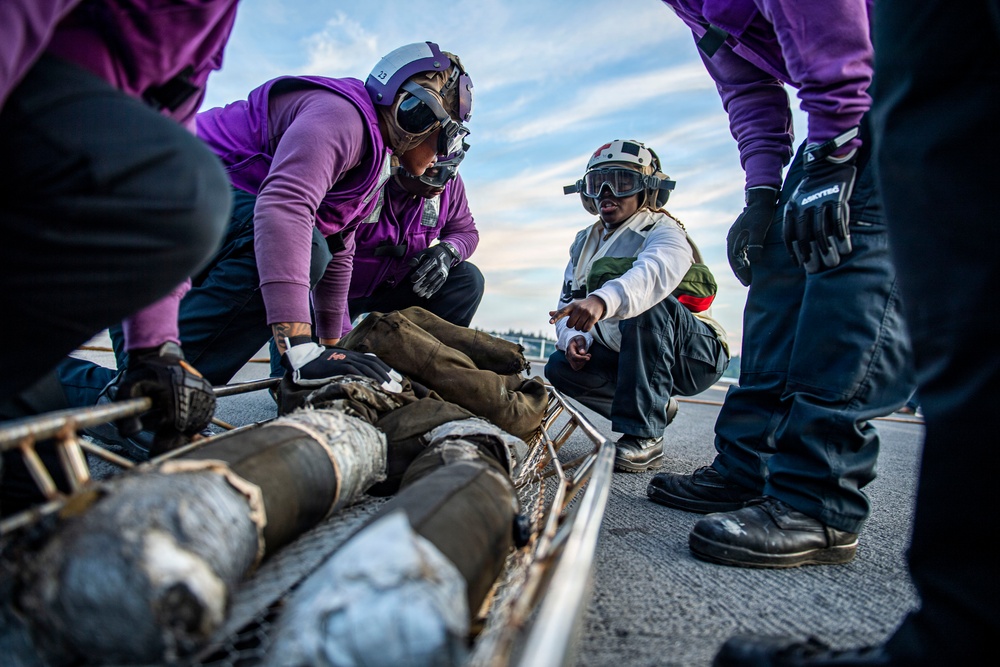 A group of military personnel wearing purple jerseys, helmets, and protective goggles are gathered on what appears to be an aircraft carrier flight deck. They are crouched around and examining what appear to be ordnance or munitions components laid out on a metal rack or cradle. One individual in a white jersey and vest appears to be supervising or directing the activity.