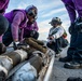A group of military personnel wearing purple jerseys, helmets, and protective goggles are gathered on what appears to be an aircraft carrier flight deck. They are crouched around and examining what appear to be ordnance or munitions components laid out on a metal rack or cradle. One individual in a white jersey and vest appears to be supervising or directing the activity.