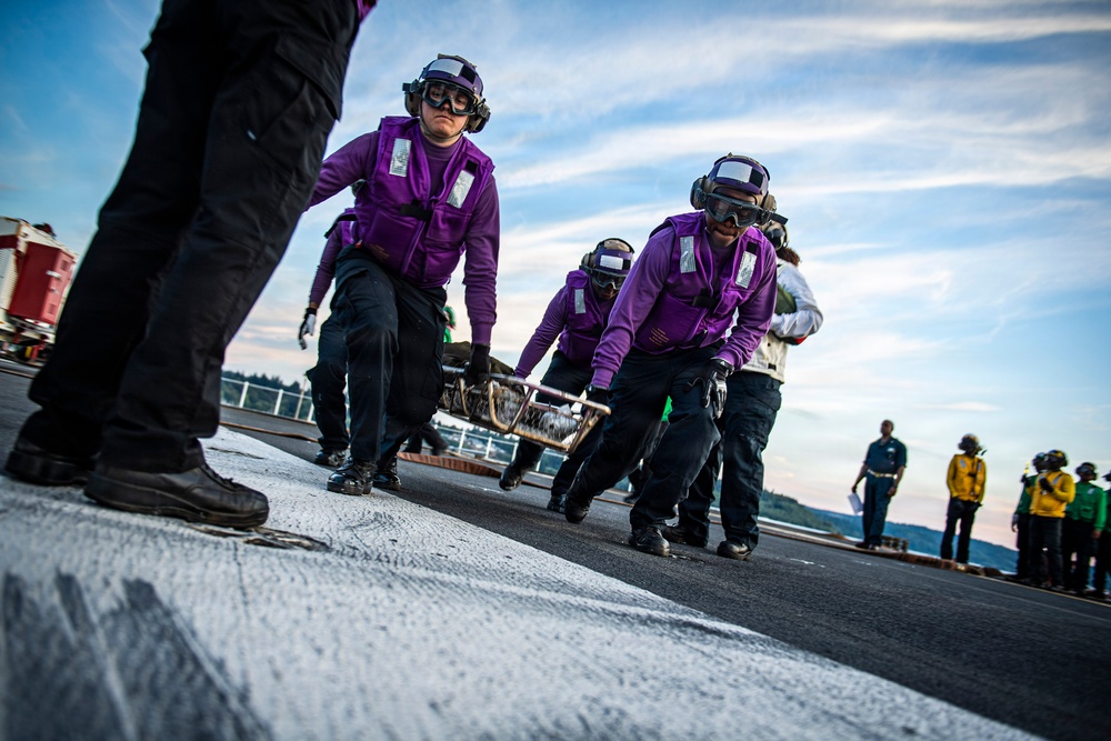 The image shows a group of U.S. Navy personnel in purple jerseys and protective gear conducting operations on an aircraft carrier flight deck. They appear to be carrying a stretcher or equipment during what looks like a drill or operational exercise. Additional crew members in yellow and green jerseys are visible in the background, indicating a multi-role flight deck operation.