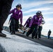 The image shows a group of U.S. Navy personnel in purple jerseys and protective gear conducting operations on an aircraft carrier flight deck. They appear to be carrying a stretcher or equipment during what looks like a drill or operational exercise. Additional crew members in yellow and green jerseys are visible in the background, indicating a multi-role flight deck operation.