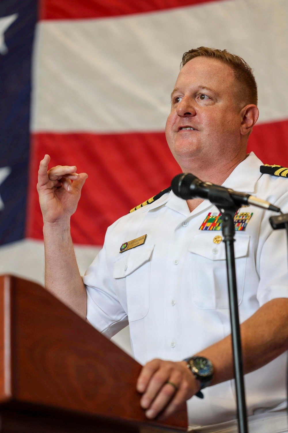 A U.S. Navy officer in white service uniform is speaking at a podium with a microphone, gesturing with his right hand. He is wearing multiple service ribbons, rank insignia consistent with a senior officer (Commander or Captain), and a name tag. An American flag is displayed prominently in the background.