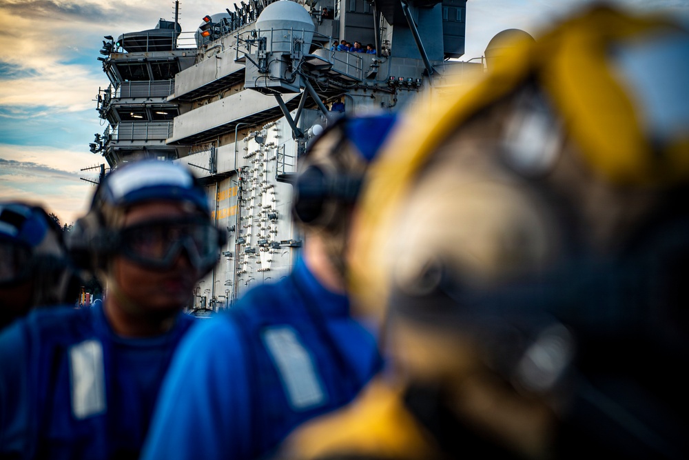 The image shows naval personnel on the flight deck of a large aircraft carrier, wearing blue jerseys, protective goggles, and hearing protection headsets. A large island superstructure dominates the background, featuring radar domes, communication equipment, and observation platforms with additional personnel visible. The foreground is intentionally blurred with shallow depth of field, focusing attention on the carrier's island structure.