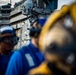 The image shows naval personnel on the flight deck of a large aircraft carrier, wearing blue jerseys, protective goggles, and hearing protection headsets. A large island superstructure dominates the background, featuring radar domes, communication equipment, and observation platforms with additional personnel visible. The foreground is intentionally blurred with shallow depth of field, focusing attention on the carrier's island structure.