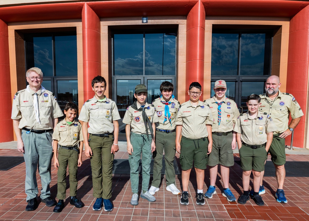 A group of Boy Scouts and two adult leaders pose for a group photo in front of a modern building with red architectural columns and large glass doors. The group consists of approximately six youth scouts and two adult leaders, all wearing BSA (Boy Scouts of America) tan uniforms with various patches and insignia. The photo appears to be taken at an official scouting event or venue.