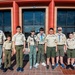 A group of Boy Scouts and two adult leaders pose for a group photo in front of a modern building with red architectural columns and large glass doors. The group consists of approximately six youth scouts and two adult leaders, all wearing BSA (Boy Scouts of America) tan uniforms with various patches and insignia. The photo appears to be taken at an official scouting event or venue.