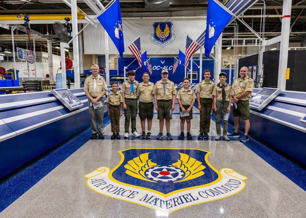 A group of Boy Scouts and two adult leaders pose for a group photo inside the Oklahoma City Air Logistics Complex (OC-ALC) facility. They are standing in front of flags including U.S. and Air Force flags, with the Air Force Materiel Command emblem prominently displayed on the floor in the foreground. The setting appears to be an official visit or tour of the military maintenance facility.