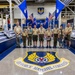 A group of Boy Scouts and two adult leaders pose for a group photo inside the Oklahoma City Air Logistics Complex (OC-ALC) facility. They are standing in front of flags including U.S. and Air Force flags, with the Air Force Materiel Command emblem prominently displayed on the floor in the foreground. The setting appears to be an official visit or tour of the military maintenance facility.