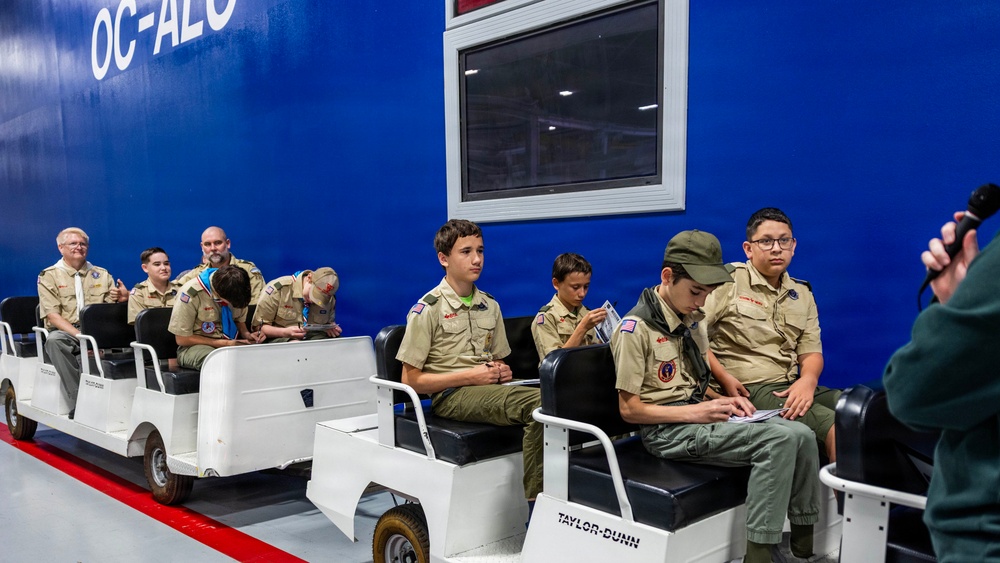 A group of Boy Scouts in tan uniforms are seated on Taylor-Dunn electric transport vehicles inside a facility with a bright blue wall marked 'OC-ALC'. The group includes several youth scouts and at least two adult leaders, appearing to be on a guided tour or educational visit. A person at the right edge of the frame appears to be recording or photographing the group.
