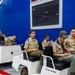 A group of Boy Scouts in tan uniforms are seated on Taylor-Dunn electric transport vehicles inside a facility with a bright blue wall marked 'OC-ALC'. The group includes several youth scouts and at least two adult leaders, appearing to be on a guided tour or educational visit. A person at the right edge of the frame appears to be recording or photographing the group.