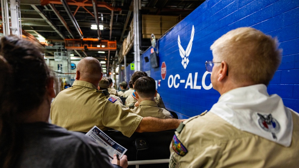 A group of personnel in tan/khaki military-style uniforms are seated in an industrial facility, facing a large blue wall bearing the U.S. Air Force symbol and the text 'OC-ALC' (Oklahoma City Air Logistics Complex). One individual is holding a document labeled 'Mission Brief.' The setting appears to be a briefing or orientation event inside a maintenance or logistics hangar.