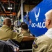 A group of personnel in tan/khaki military-style uniforms are seated in an industrial facility, facing a large blue wall bearing the U.S. Air Force symbol and the text 'OC-ALC' (Oklahoma City Air Logistics Complex). One individual is holding a document labeled 'Mission Brief.' The setting appears to be a briefing or orientation event inside a maintenance or logistics hangar.