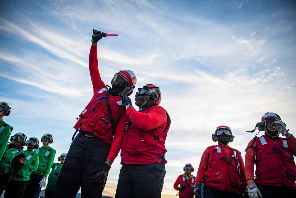 The image shows military personnel, likely U.S. Navy sailors, on what appears to be an aircraft carrier flight deck. Personnel are wearing color-coded jerseys — red and green — along with helmets, goggles, and hearing protection, consistent with carrier flight deck operations. One individual in red is holding a lighted wand/baton raised above their head, suggesting active signaling or marshaling activity.