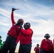 The image shows military personnel, likely U.S. Navy sailors, on what appears to be an aircraft carrier flight deck. Personnel are wearing color-coded jerseys — red and green — along with helmets, goggles, and hearing protection, consistent with carrier flight deck operations. One individual in red is holding a lighted wand/baton raised above their head, suggesting active signaling or marshaling activity.