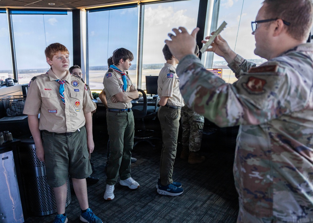 A group of Boy Scouts in tan uniforms with neckerchiefs and merit badge sashes are receiving a tour of what appears to be a military air traffic control tower. A military personnel member in MultiCam/OCP camouflage uniform is briefing or instructing the scouts, holding what appears to be printed documents or charts. The large windows in the background reveal an airfield or airport tarmac.
