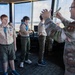 A group of Boy Scouts in tan uniforms with neckerchiefs and merit badge sashes are receiving a tour of what appears to be a military air traffic control tower. A military personnel member in MultiCam/OCP camouflage uniform is briefing or instructing the scouts, holding what appears to be printed documents or charts. The large windows in the background reveal an airfield or airport tarmac.