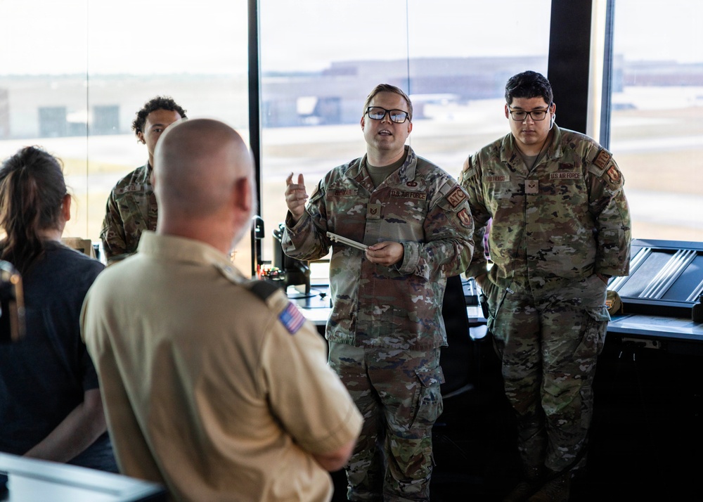 Several U.S. Air Force personnel in Operational Camouflage Pattern (OCP) uniforms are gathered in what appears to be an air traffic control tower, indicated by the large windows overlooking an airfield. One Airman wearing glasses appears to be briefing or presenting to a group that includes a person in a tan/khaki uniform facing away from the camera. The setting suggests a training, orientation, or inspection event.