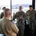 Several U.S. Air Force personnel in Operational Camouflage Pattern (OCP) uniforms are gathered in what appears to be an air traffic control tower, indicated by the large windows overlooking an airfield. One Airman wearing glasses appears to be briefing or presenting to a group that includes a person in a tan/khaki uniform facing away from the camera. The setting suggests a training, orientation, or inspection event.