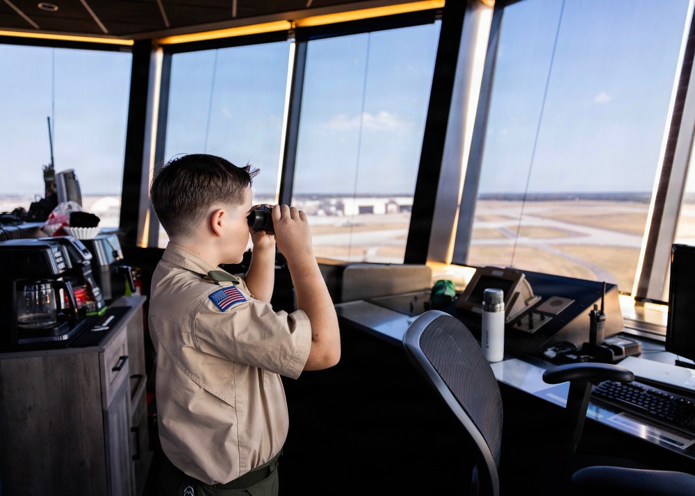 A young person wearing a Boy Scouts of America uniform with a US flag patch is looking through binoculars from inside an air traffic control tower. The tower cab overlooks an airport tarmac and runway area. The setting suggests this is likely an educational or tour visit to the facility.