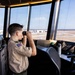 A young person wearing a Boy Scouts of America uniform with a US flag patch is looking through binoculars from inside an air traffic control tower. The tower cab overlooks an airport tarmac and runway area. The setting suggests this is likely an educational or tour visit to the facility.