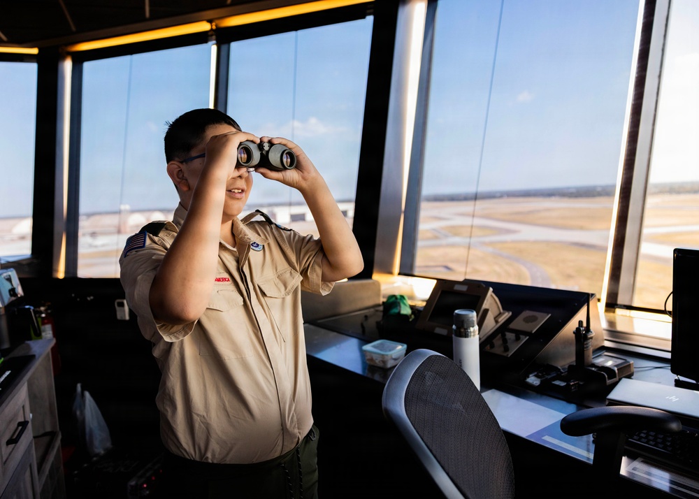 A young person wearing a Boy Scouts of America uniform is standing in an airport control tower, looking through binoculars toward the airfield. The setting appears to be an educational or tour visit, with the child exploring the tower's equipment and view. Airport runways and taxiways are visible through the large panoramic windows behind them.