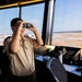 A young person wearing a Boy Scouts of America uniform is standing in an airport control tower, looking through binoculars toward the airfield. The setting appears to be an educational or tour visit, with the child exploring the tower's equipment and view. Airport runways and taxiways are visible through the large panoramic windows behind them.