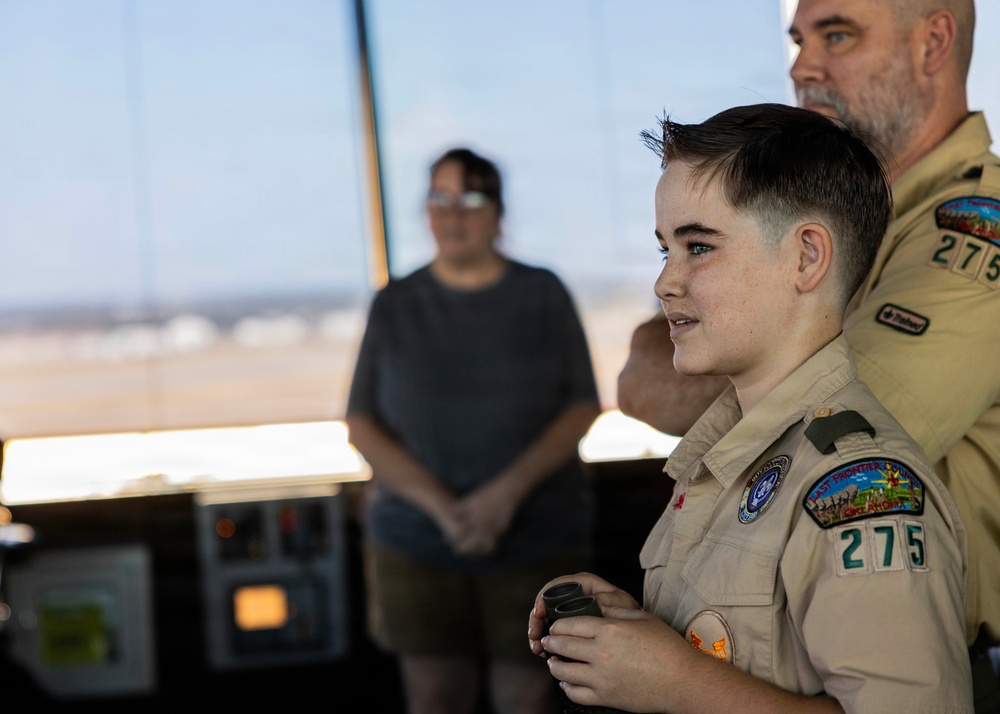 A young Boy Scout wearing a tan uniform with troop number 275 and 'Last Frontier' council patches stands in what appears to be an airport or military air traffic control tower. An adult Scout leader in a matching uniform stands behind the youth, and a civilian observer stands in the background. The setting suggests an educational or field trip visit to an aviation facility.