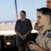 A young Boy Scout wearing a tan uniform with troop number 275 and 'Last Frontier' council patches stands in what appears to be an airport or military air traffic control tower. An adult Scout leader in a matching uniform stands behind the youth, and a civilian observer stands in the background. The setting suggests an educational or field trip visit to an aviation facility.
