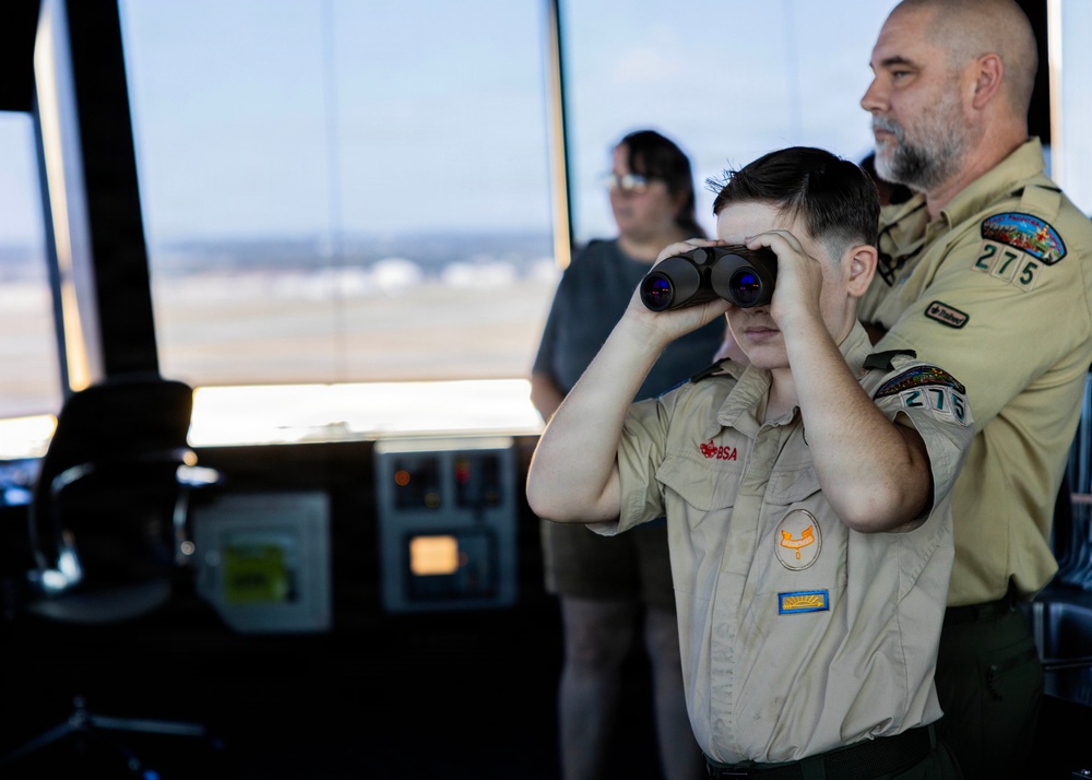 A Boy Scout (BSA) youth member looks through binoculars while standing in what appears to be an airport or airfield control tower. An adult Scout leader wearing a 'Troop 275' patch stands beside him, and a third person is visible in the background. The setting suggests a guided educational visit to an air traffic control facility.