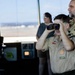 A Boy Scout (BSA) youth member looks through binoculars while standing in what appears to be an airport or airfield control tower. An adult Scout leader wearing a 'Troop 275' patch stands beside him, and a third person is visible in the background. The setting suggests a guided educational visit to an air traffic control facility.