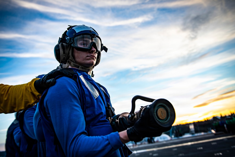 A naval flight deck crew member wearing a blue jersey, flight deck helmet, ear protection, and protective goggles is holding a fuel nozzle or hose coupling on what appears to be an aircraft carrier flight deck. The image is taken at low angle during sunset or sunrise, giving a dramatic sky backdrop with warm orange and blue tones. A second crew member in a yellow glove is partially visible on the left side of the frame.