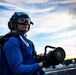 A naval flight deck crew member wearing a blue jersey, flight deck helmet, ear protection, and protective goggles is holding a fuel nozzle or hose coupling on what appears to be an aircraft carrier flight deck. The image is taken at low angle during sunset or sunrise, giving a dramatic sky backdrop with warm orange and blue tones. A second crew member in a yellow glove is partially visible on the left side of the frame.