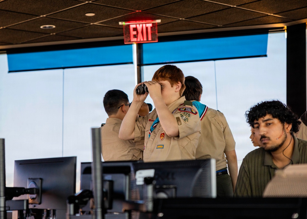 A group of Boy Scouts are visiting what appears to be an air traffic control tower, as evidenced by the large panoramic windows, computer workstations, and professional equipment. A red-haired scout in full uniform with merit badge sash is looking through binoculars toward the windows, while other scouts and at least one staff member are present. The setting suggests an educational tour of an aviation facility.