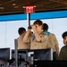 A group of Boy Scouts are visiting what appears to be an air traffic control tower, as evidenced by the large panoramic windows, computer workstations, and professional equipment. A red-haired scout in full uniform with merit badge sash is looking through binoculars toward the windows, while other scouts and at least one staff member are present. The setting suggests an educational tour of an aviation facility.