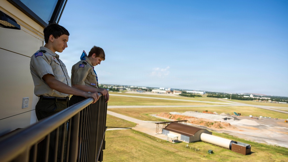 Two young people wearing Boy Scout uniforms stand on an elevated outdoor balcony or observation deck, looking down over an airport or airfield facility. The view behind them shows runways, taxiways, open grassy areas, and what appears to be construction or maintenance activity. The setting suggests a guided tour or educational visit to an air traffic control tower or similar aviation facility.