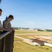 Two young people wearing Boy Scout uniforms stand on an elevated outdoor balcony or observation deck, looking down over an airport or airfield facility. The view behind them shows runways, taxiways, open grassy areas, and what appears to be construction or maintenance activity. The setting suggests a guided tour or educational visit to an air traffic control tower or similar aviation facility.