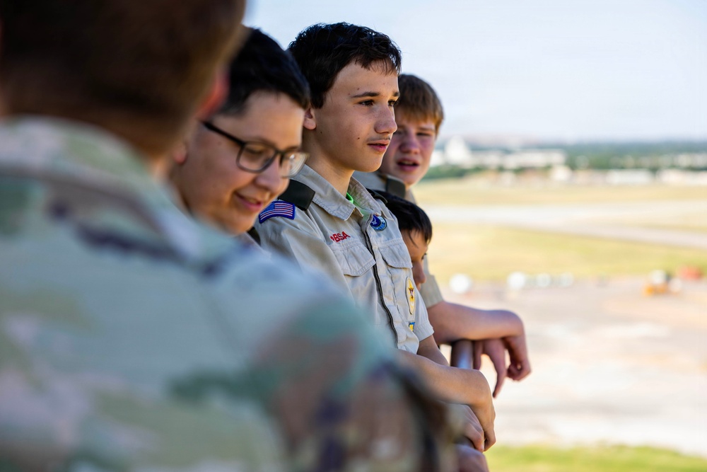 A group of young teenagers, several wearing Boy Scouts of America (BSA) uniforms, are standing at a railing overlooking what appears to be an airport tarmac or airfield. The scouts appear to be on an organized field trip or educational visit, observing aircraft operations from an elevated observation area. The image is taken during golden hour lighting, giving it a warm tone.