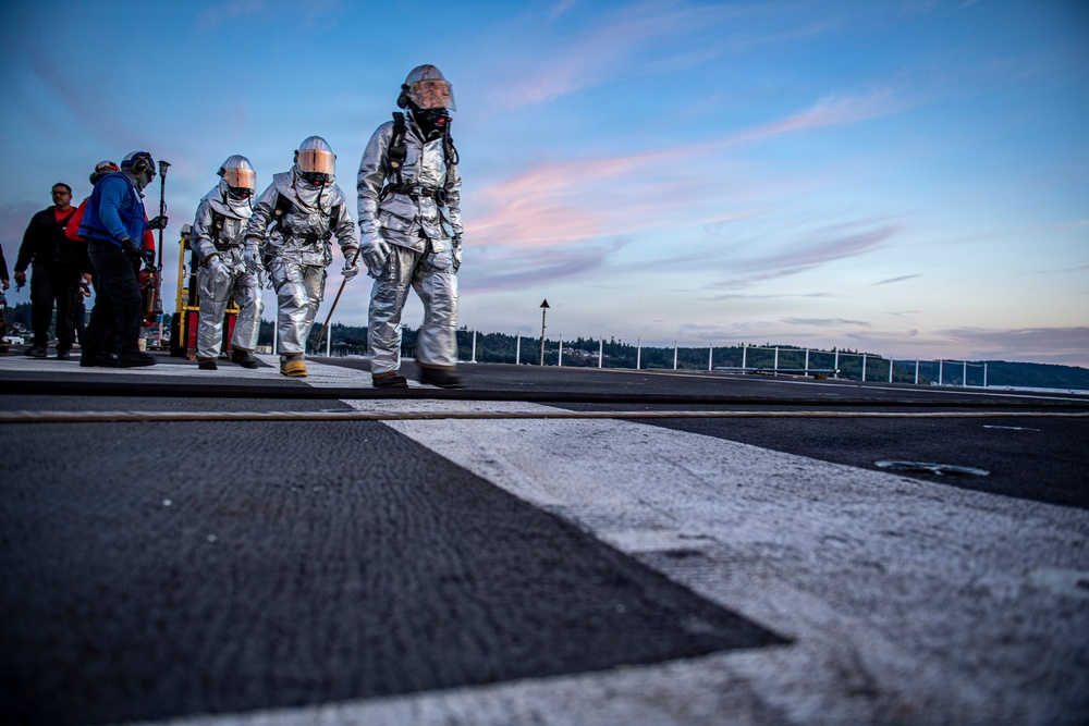 Three personnel wearing silver aluminized proximity fire suits and respirator masks walk across what appears to be a naval vessel's flight deck at dusk. Additional personnel in standard work attire are visible in the background along with firefighting equipment. The setting appears to be a military exercise or drill conducted on a ship deck near a forested coastline.