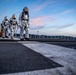 Three personnel wearing silver aluminized proximity fire suits and respirator masks walk across what appears to be a naval vessel's flight deck at dusk. Additional personnel in standard work attire are visible in the background along with firefighting equipment. The setting appears to be a military exercise or drill conducted on a ship deck near a forested coastline.