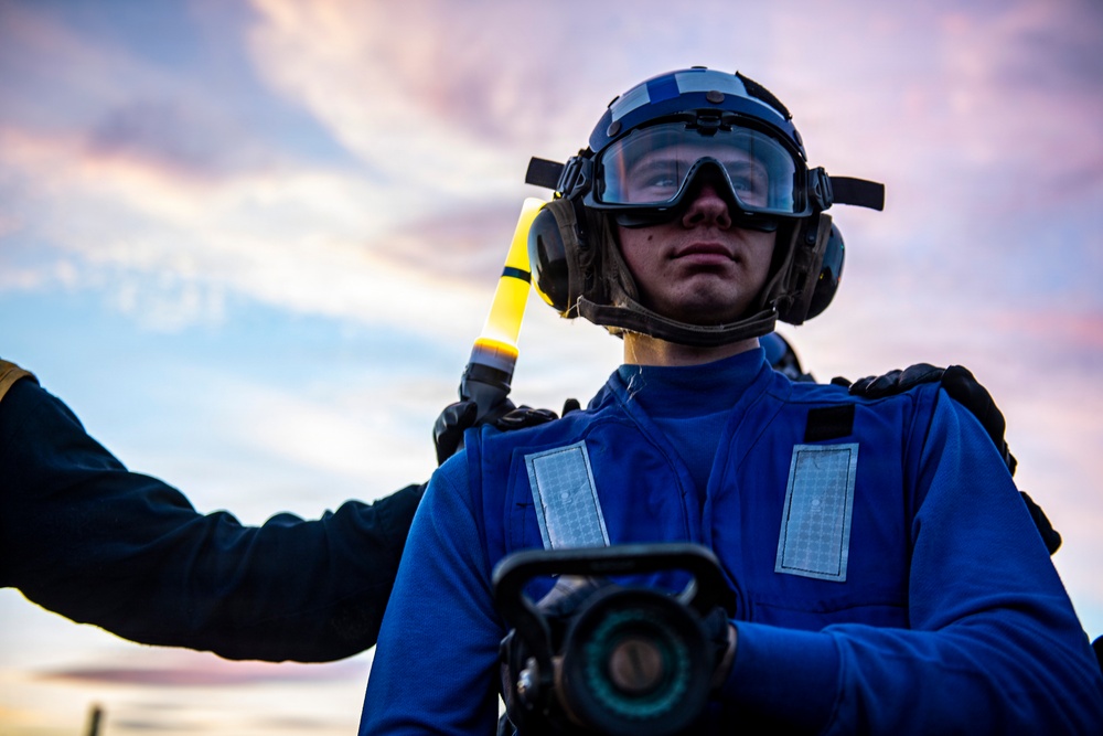 A military or naval aviation deck crew member is photographed from a low angle against a dramatic twilight sky. The individual is wearing blue flight deck gear and holding a yellow illuminated wand, consistent with aircraft handling or marshaling duties. The composition and equipment suggest this is taken aboard a naval vessel flight deck during low-light operations.