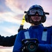 A military or naval aviation deck crew member is photographed from a low angle against a dramatic twilight sky. The individual is wearing blue flight deck gear and holding a yellow illuminated wand, consistent with aircraft handling or marshaling duties. The composition and equipment suggest this is taken aboard a naval vessel flight deck during low-light operations.